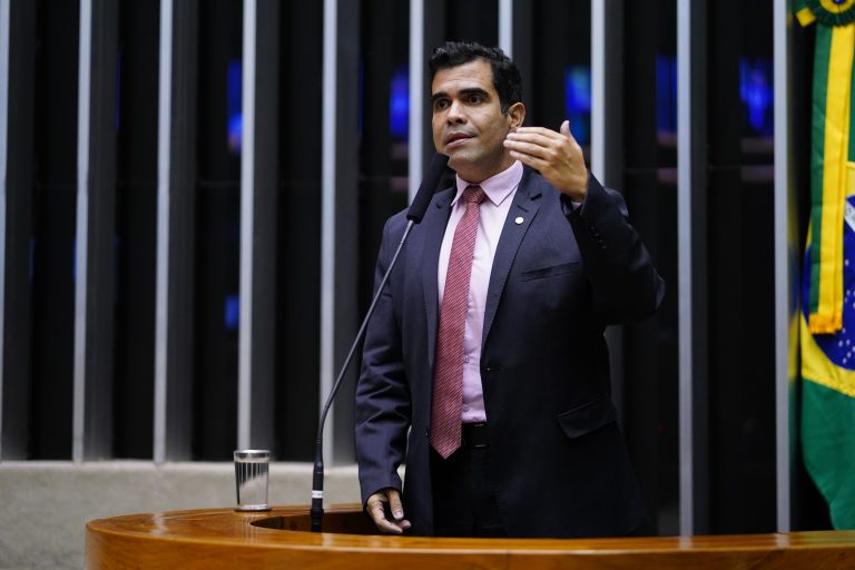 Deputado Ricardo Ayres fala ao microfone. Ele é claro, tem cabelo escuro e veste um terno azul-marinho