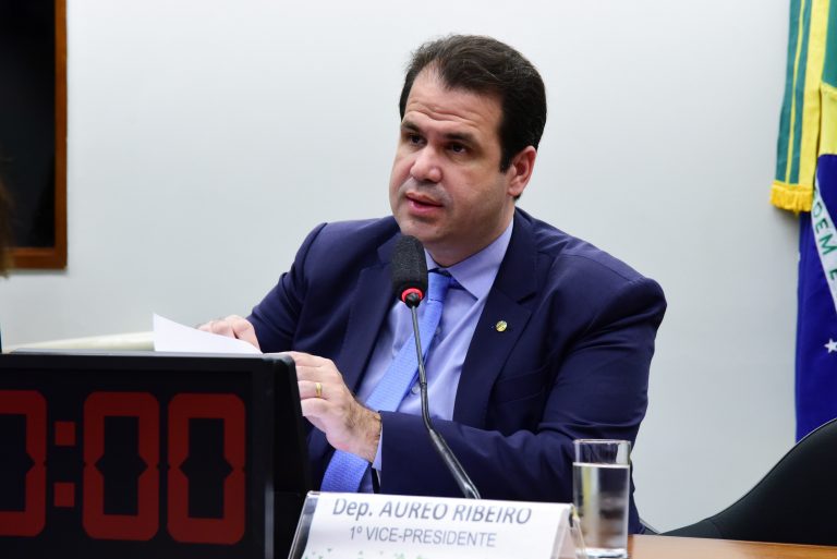 Deputado Aureo Ribeiro fala ao microfone. Ele é um homem claro e usa um terno azul marinho